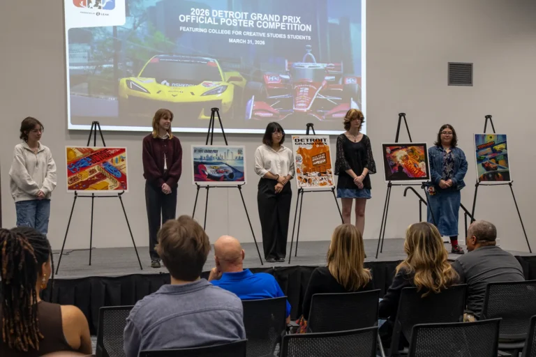 Finalists for the annual Detroit Grand Prix Poster Competition stand on stage at the College for Creative Studies next to their final poster designs for the 2026 Detroit Grand Prix