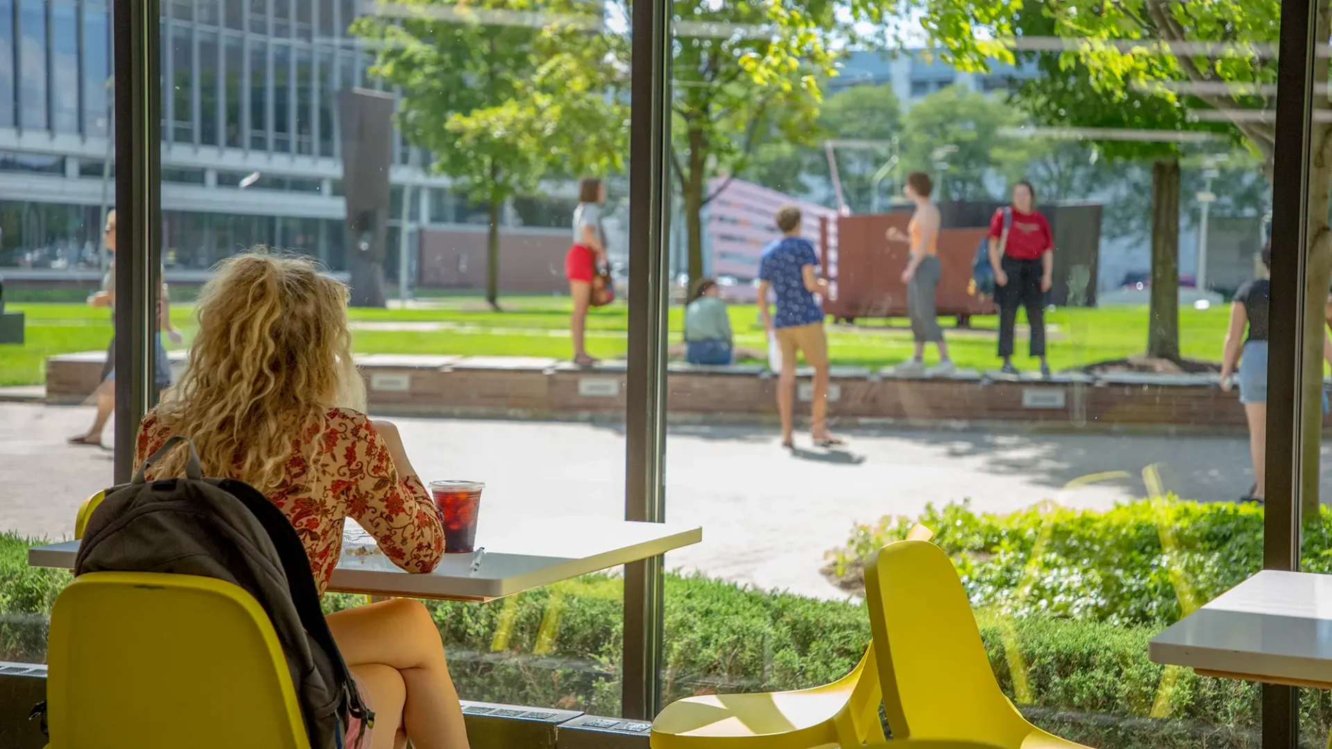 A student in the cafe looking out through the glass windows onto other students in a group in the sculpture garden on the Ford Campus