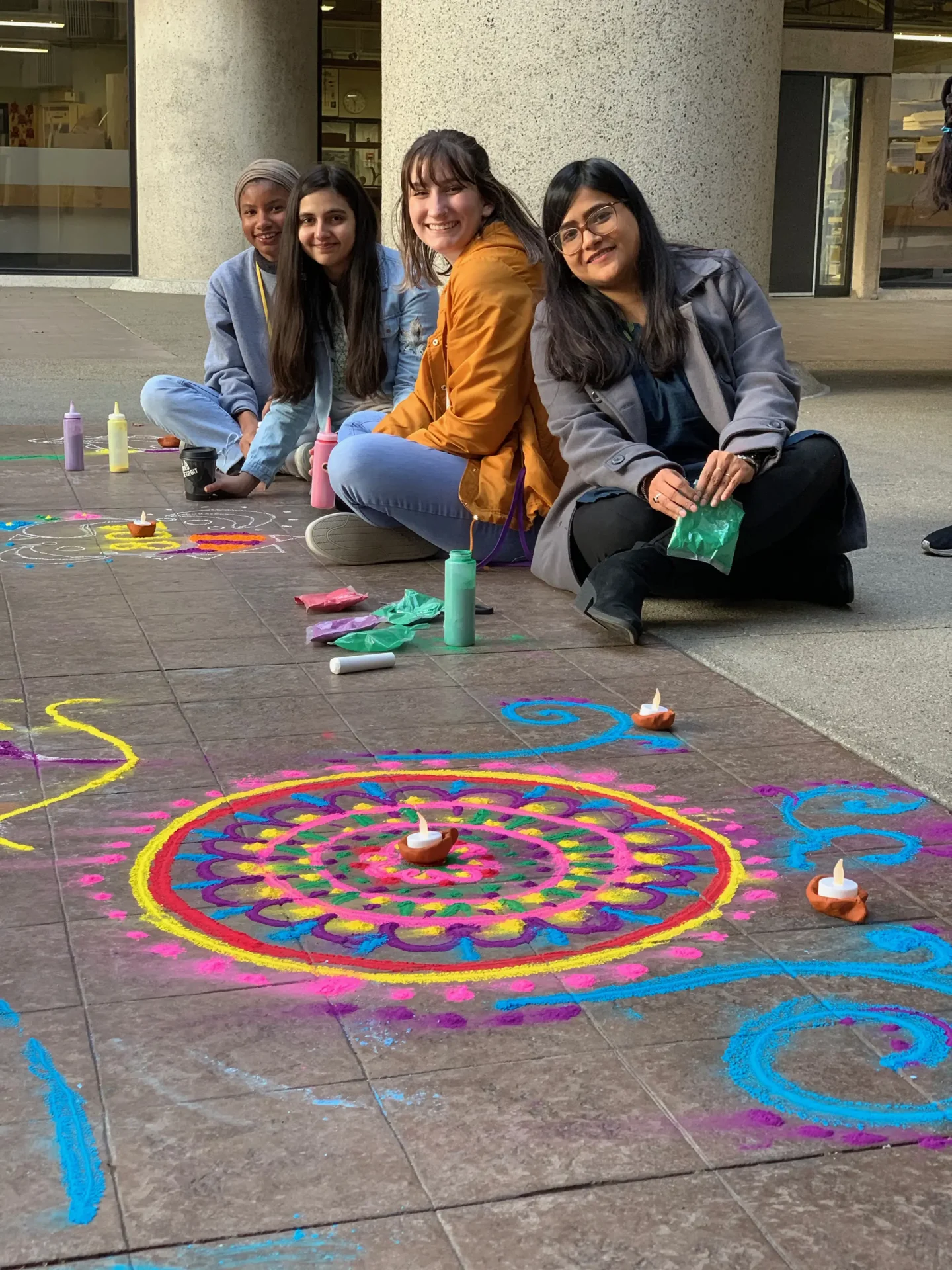 A group of students sitting on the ground next to chalk and sand artwork created for Diwali