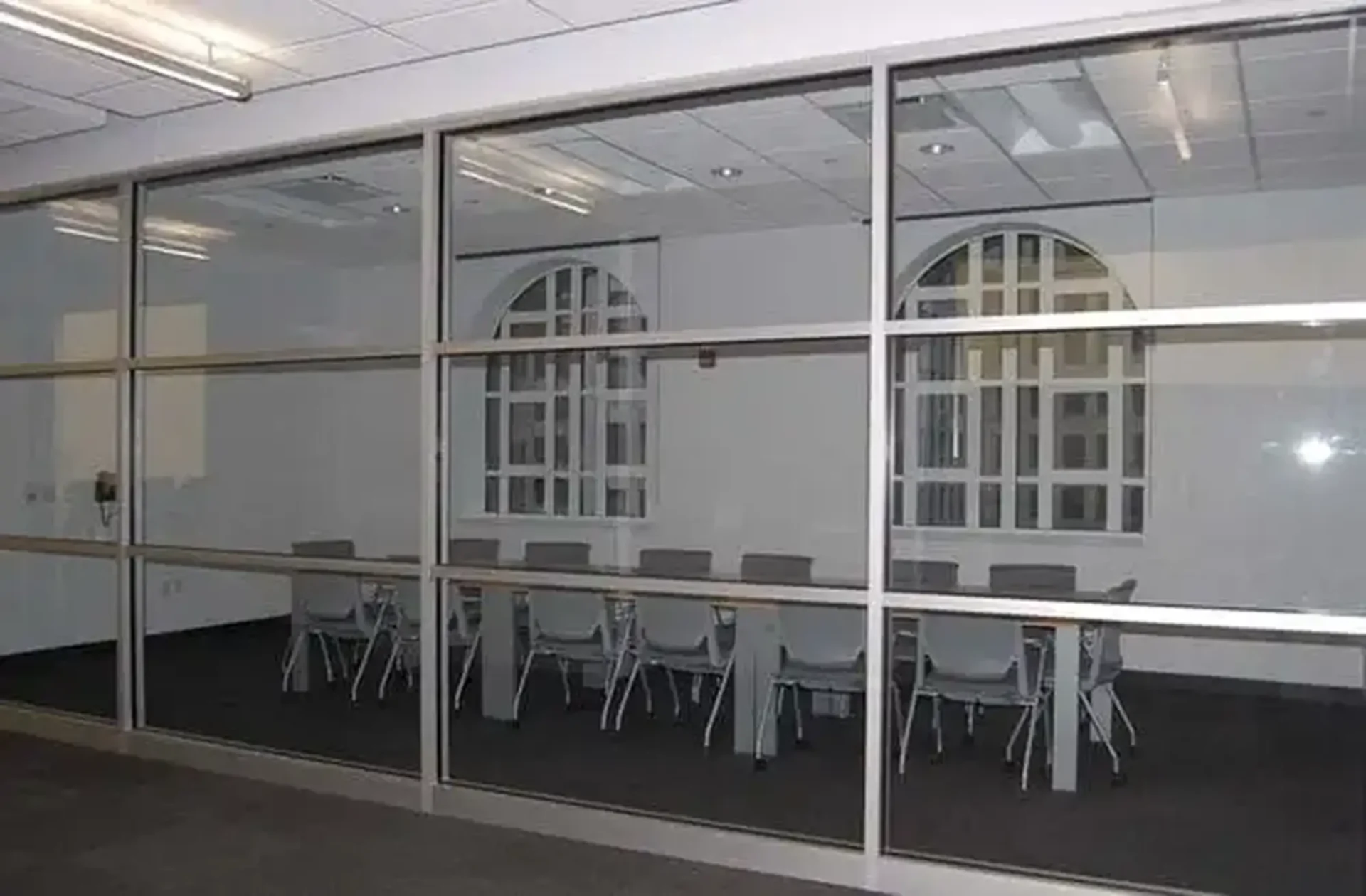 A meeting room at the Benson & Edith Ford Conference Center seen through a glass partition, featuring a long conference table and gray office chairs positioned in front of two large, distinctive arched windows with white geometric frames.