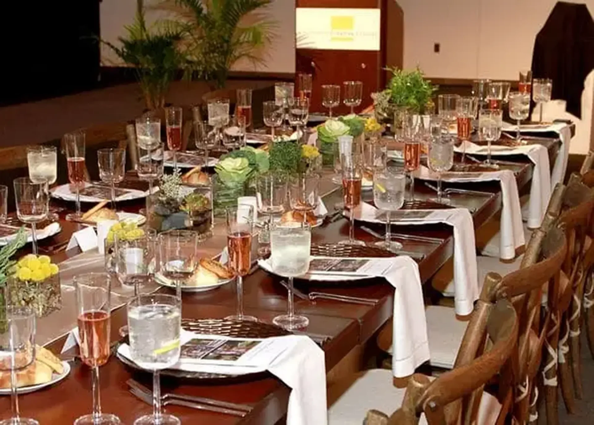 A formal, long wood dining table fully set with glassware, napkins, and floral centerpieces inside the General Motors Auditorium.