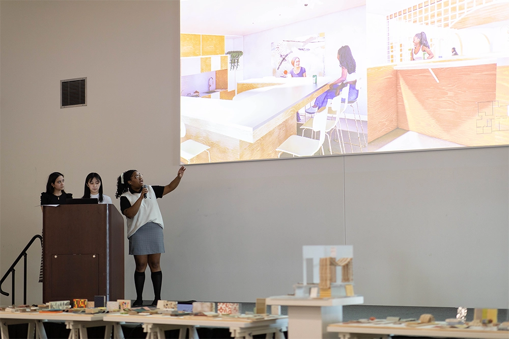 Three students present designs of a new facility to the C.L.A.S.S. Agency on a large screen at a podium with material trays in the foreground.