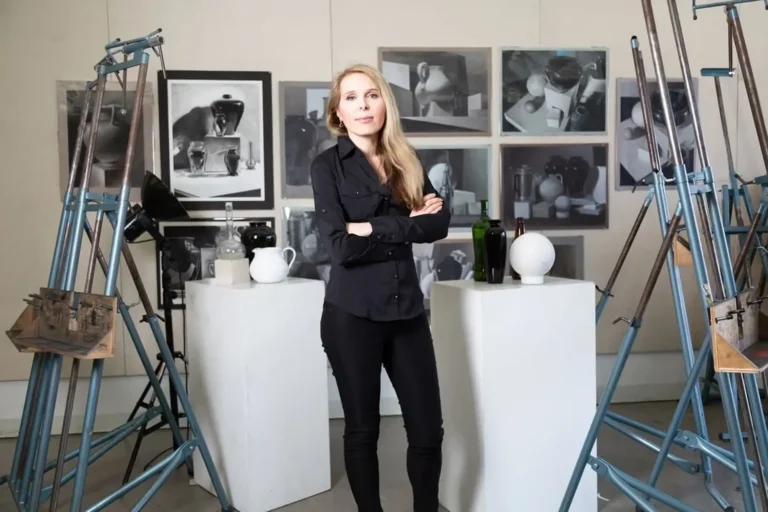 Elena McCann-Arnaoutova standing with arms crossed in an art studio, surrounded by easels, pedestals with still life objects, and framed black-and-white artwork on the wall.