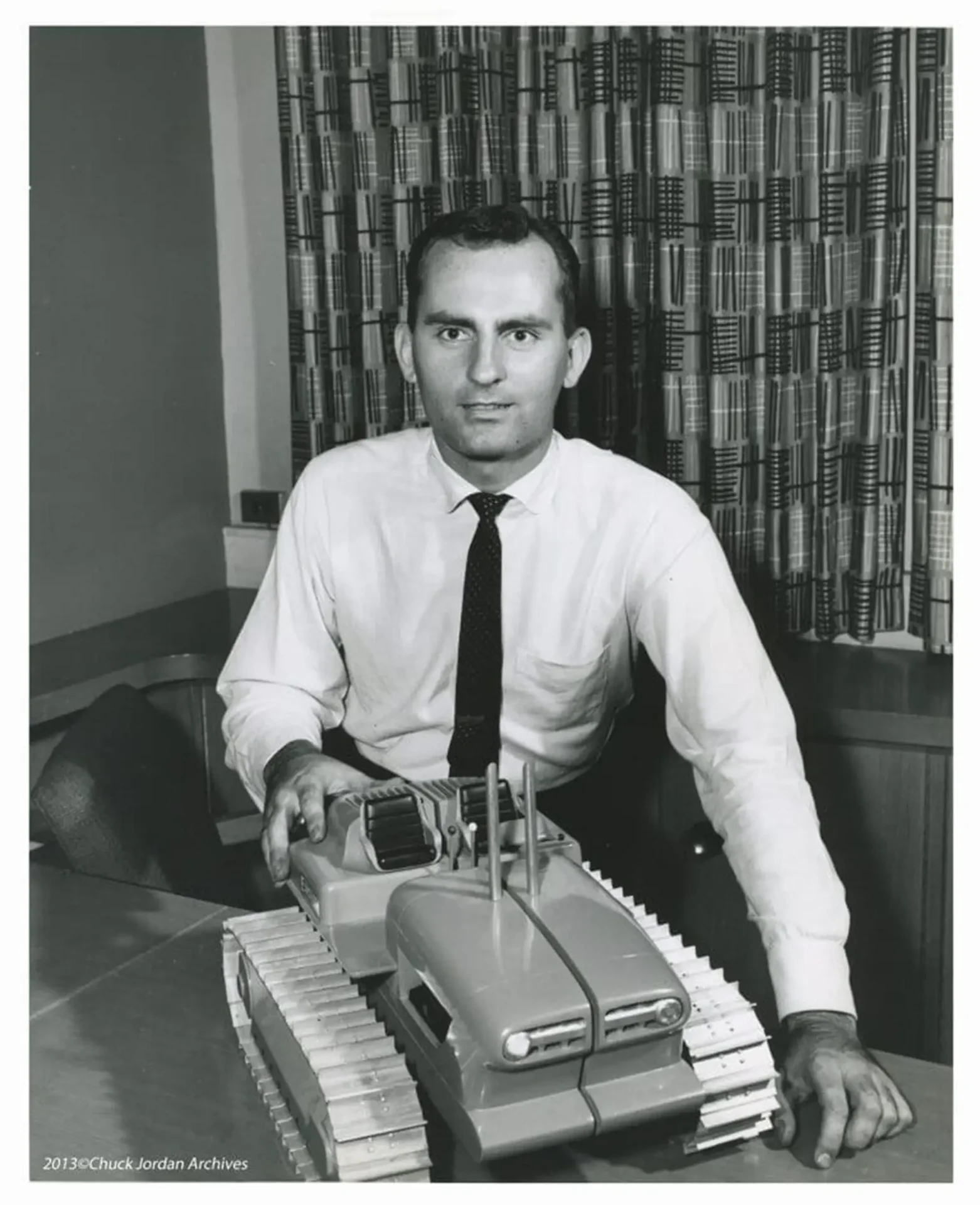 Chuck Jordan posing with an image of a model vehicle with large treads on it