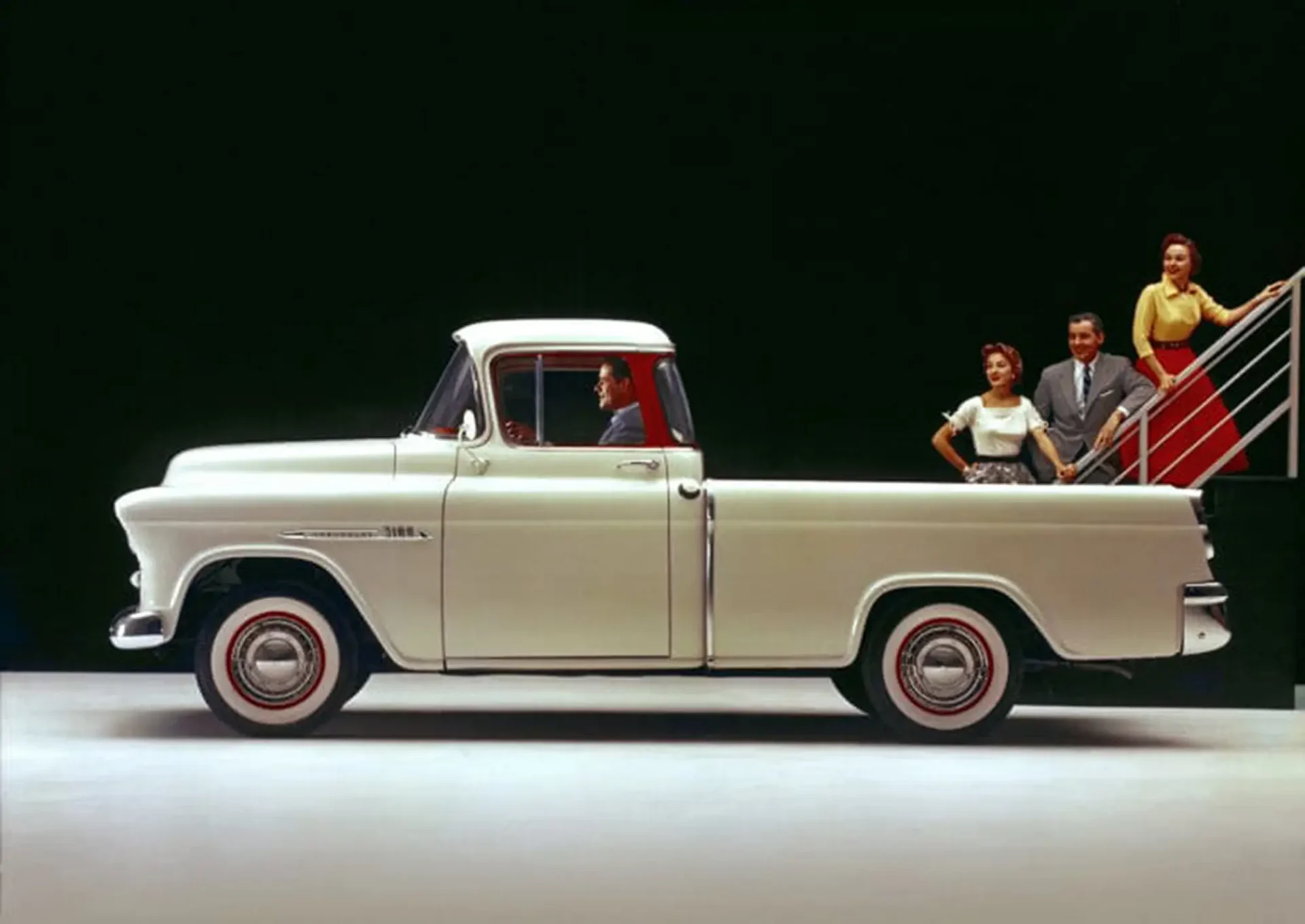 A pickup truck with a ladder coming out the back. A group of 3 people are looking at the cab of the truck as they move up the stairs