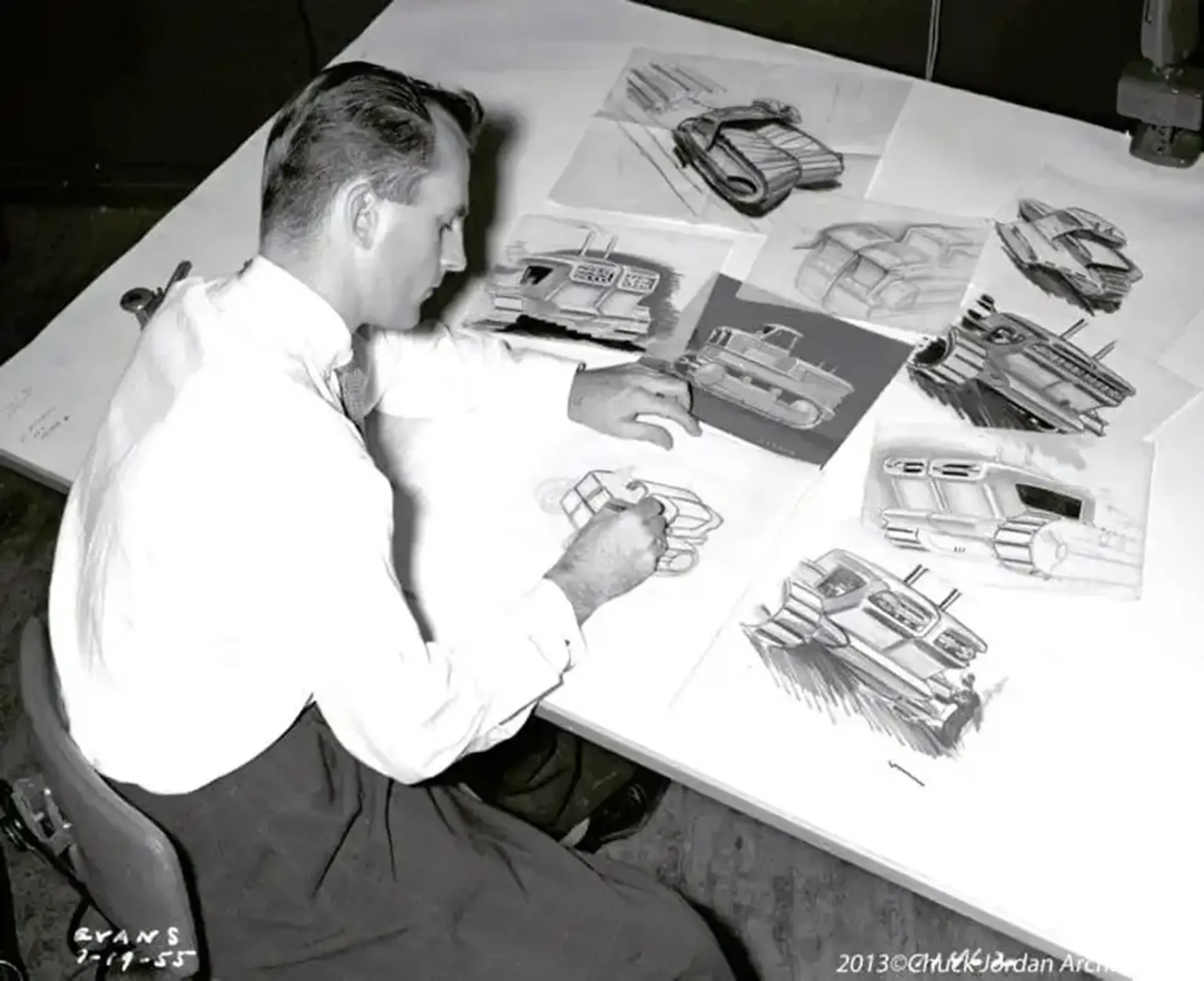 A photo of Chuck Jordan sketching at a table. The table has various other sketches scattered across it