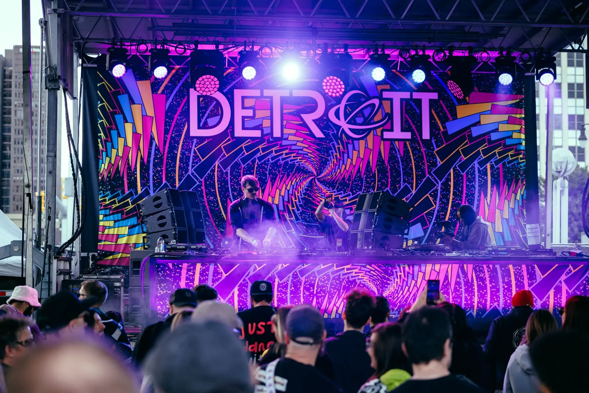 A DJ plays in front of a large crowd. There is a technicolored backdrop behind him that says "Detroit"