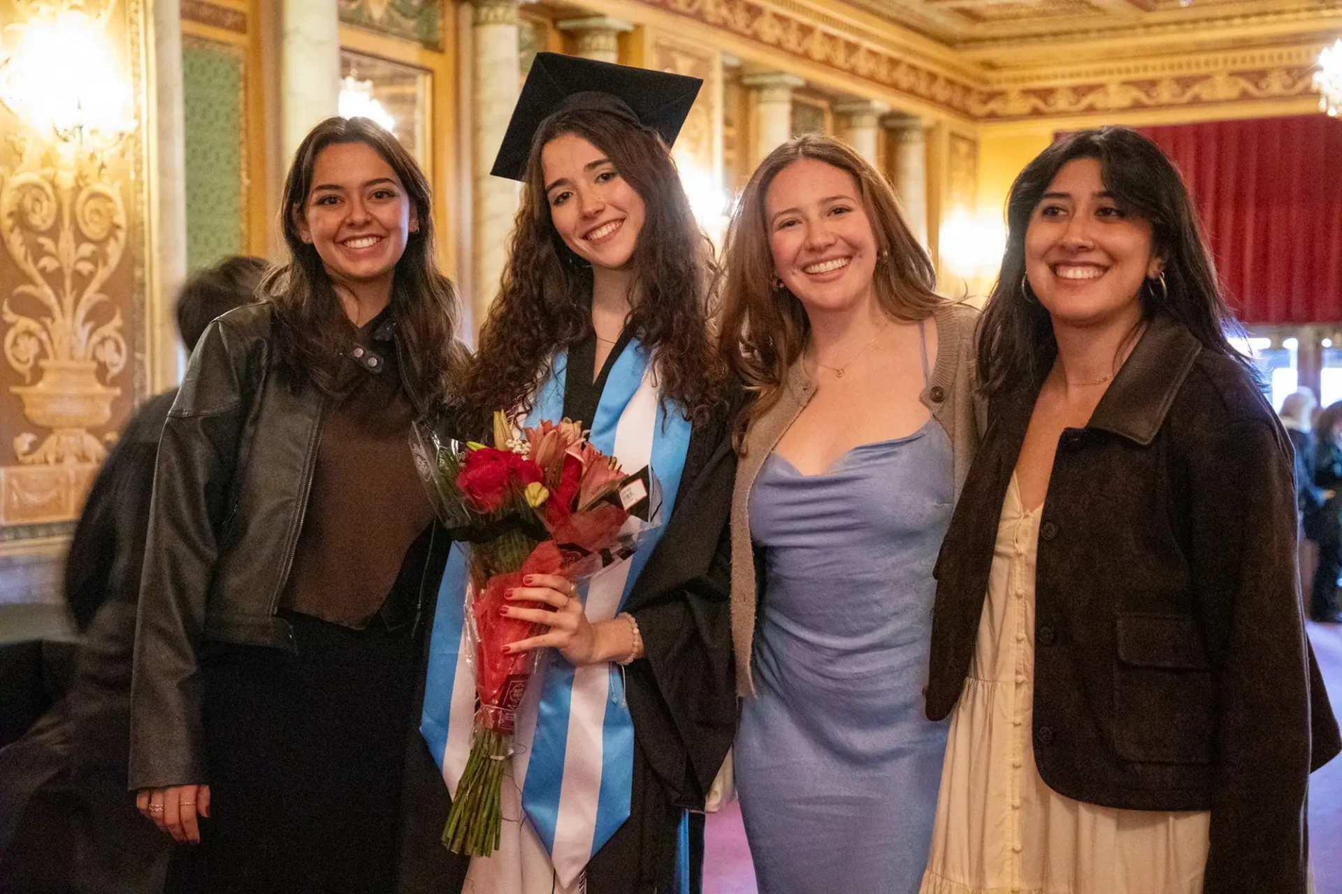 A group of girls posing for a picture at the Filmore. One of the girls is wearing her cap and holding a bouquet of flowers