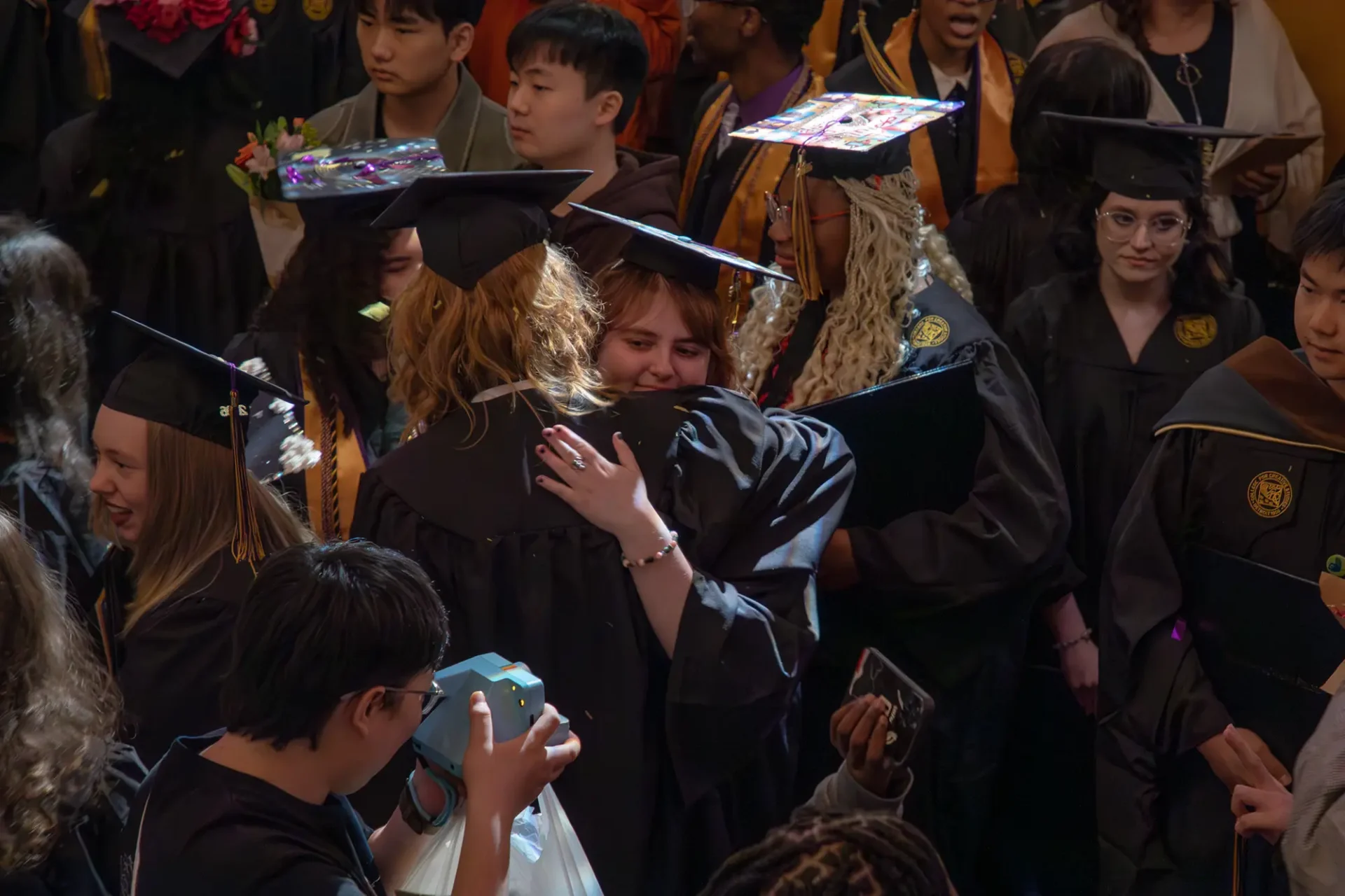 Two students are smiling and hugging eachother in crowd of graduates