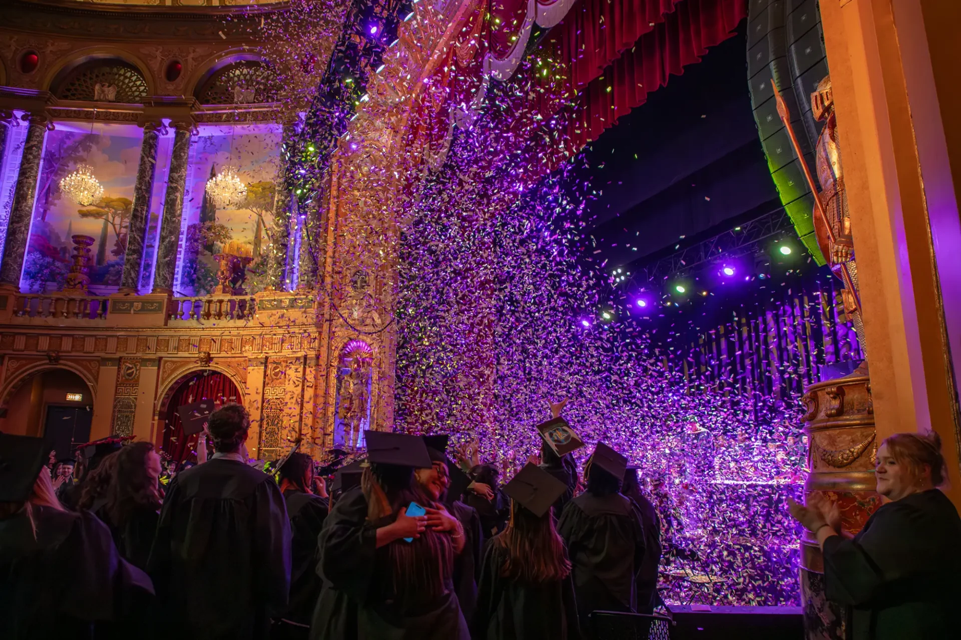 Two students hug in a group of others as brightly colored confetti falls from the ceiling