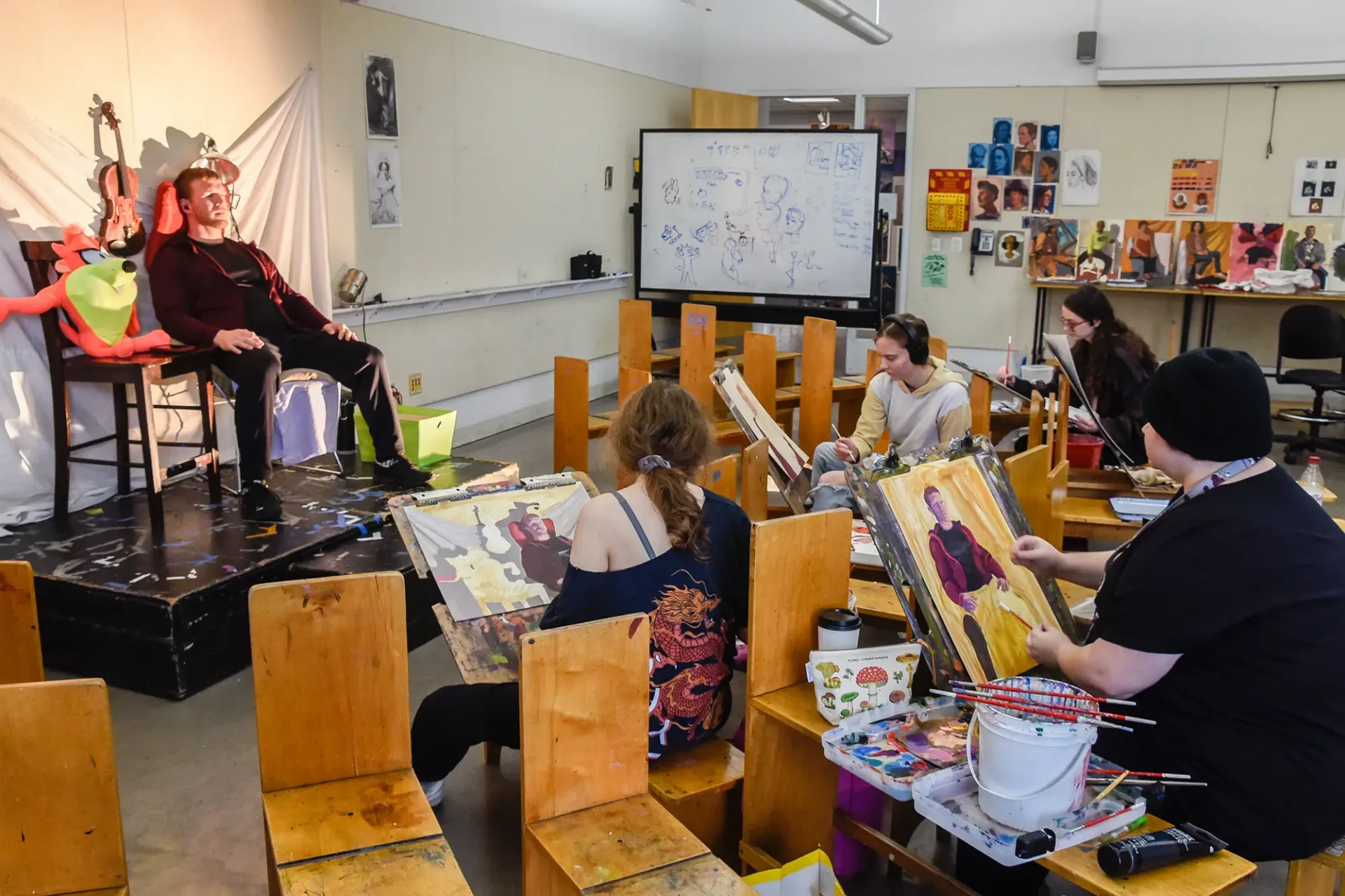 Students painting a still life of a model in a classroom.