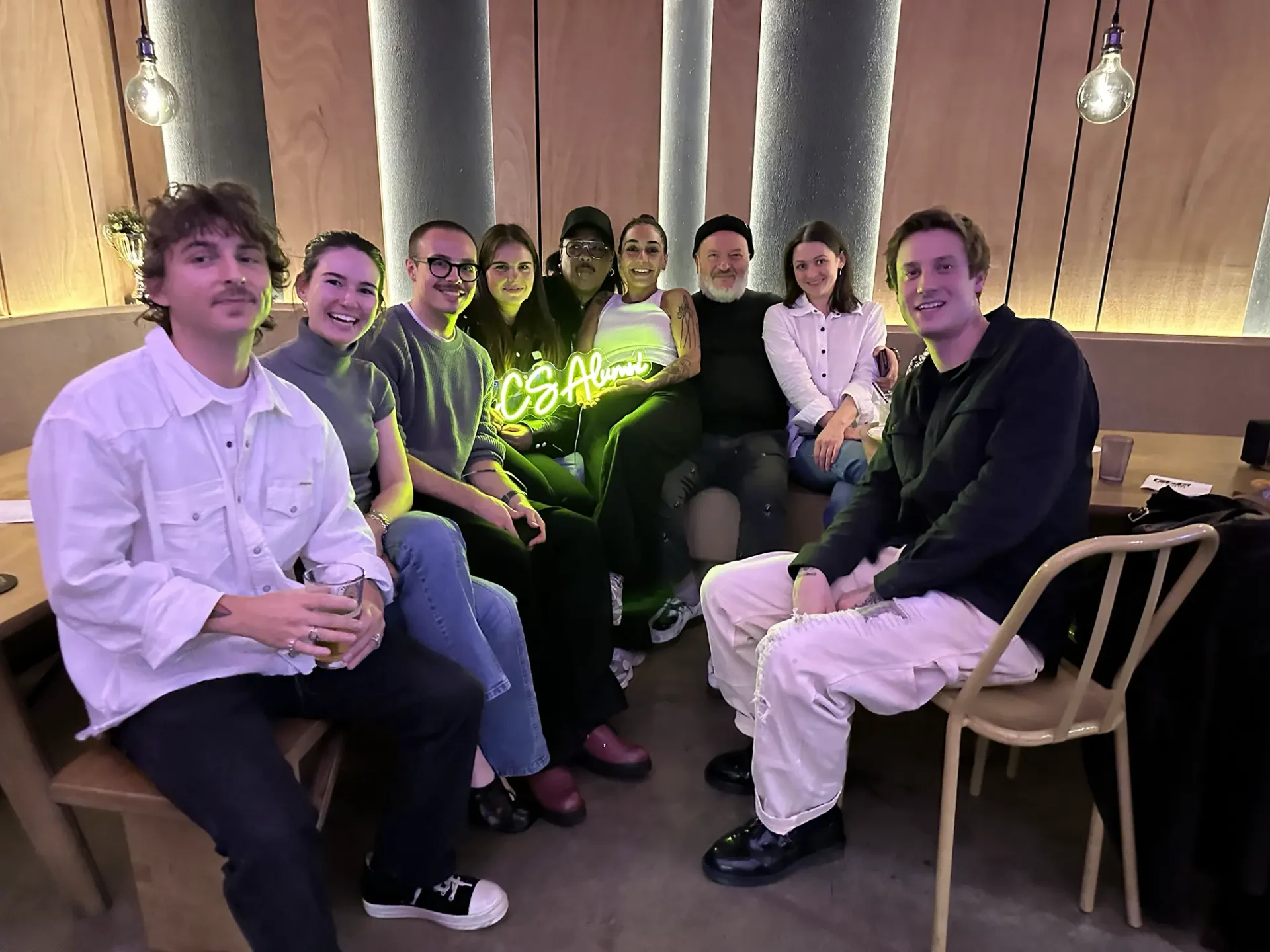 a group of alumni gathered in a bar in Brooklyn holding a yellow neon sign that reads "CCS Alumni"