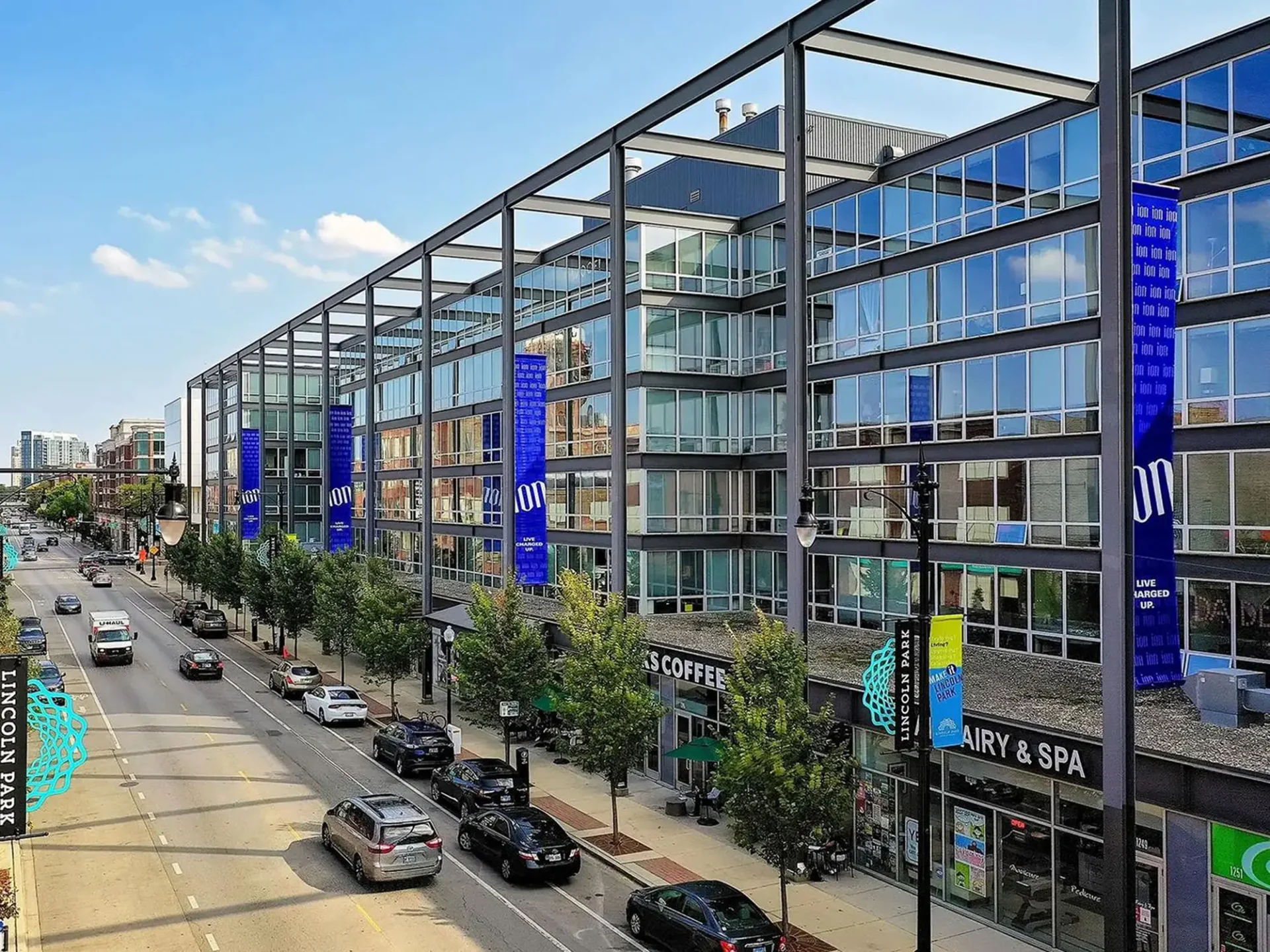 Modern multi-story building along a city street with blue banners, glass windows, and shops at ground level.