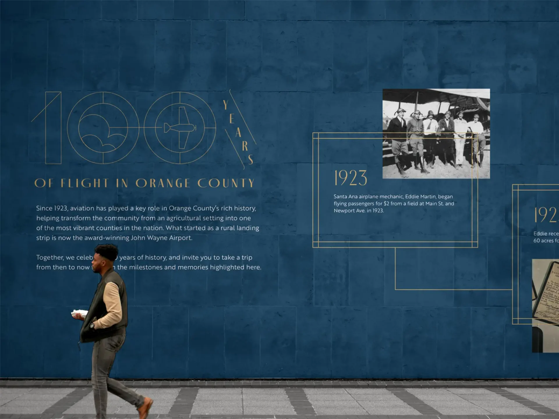 Man walking past a dark blue wall display with gold text reading "100 Years of Flight in Orange County."