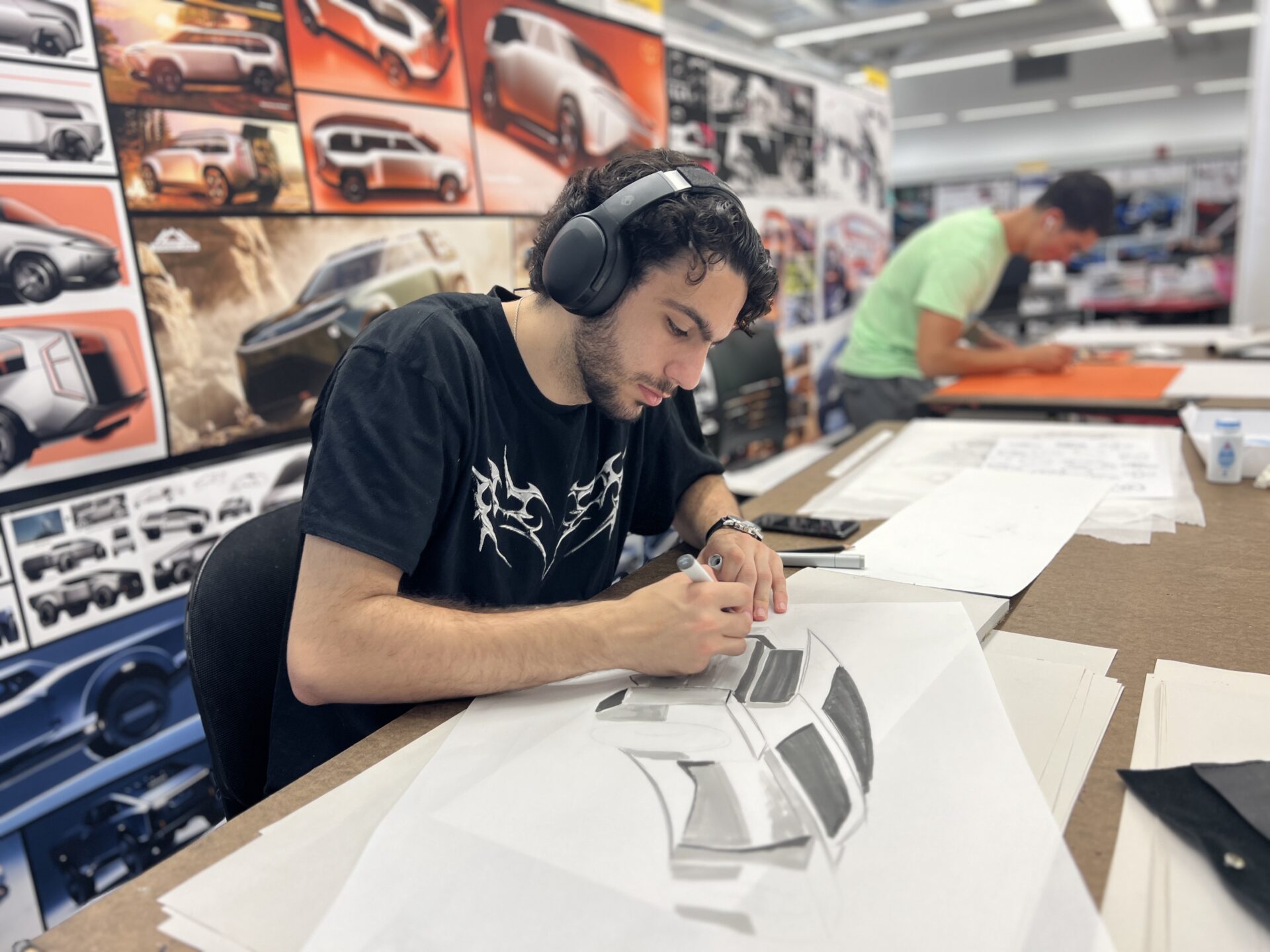 A design student wearing over-ear headphones sketching a futuristic car concept with a marker at a studio drafting table.