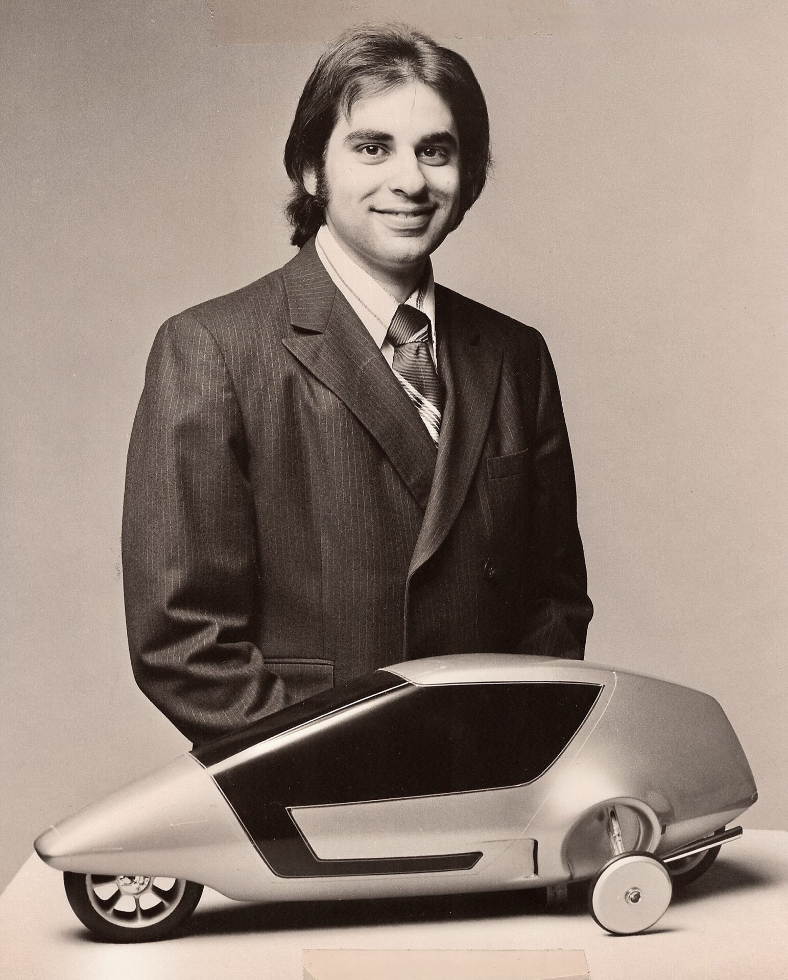 Black-and-white portrait of a young John Manoogian smiling in a suit, standing behind a sleek, futuristic silver model car with dark tinted windows.