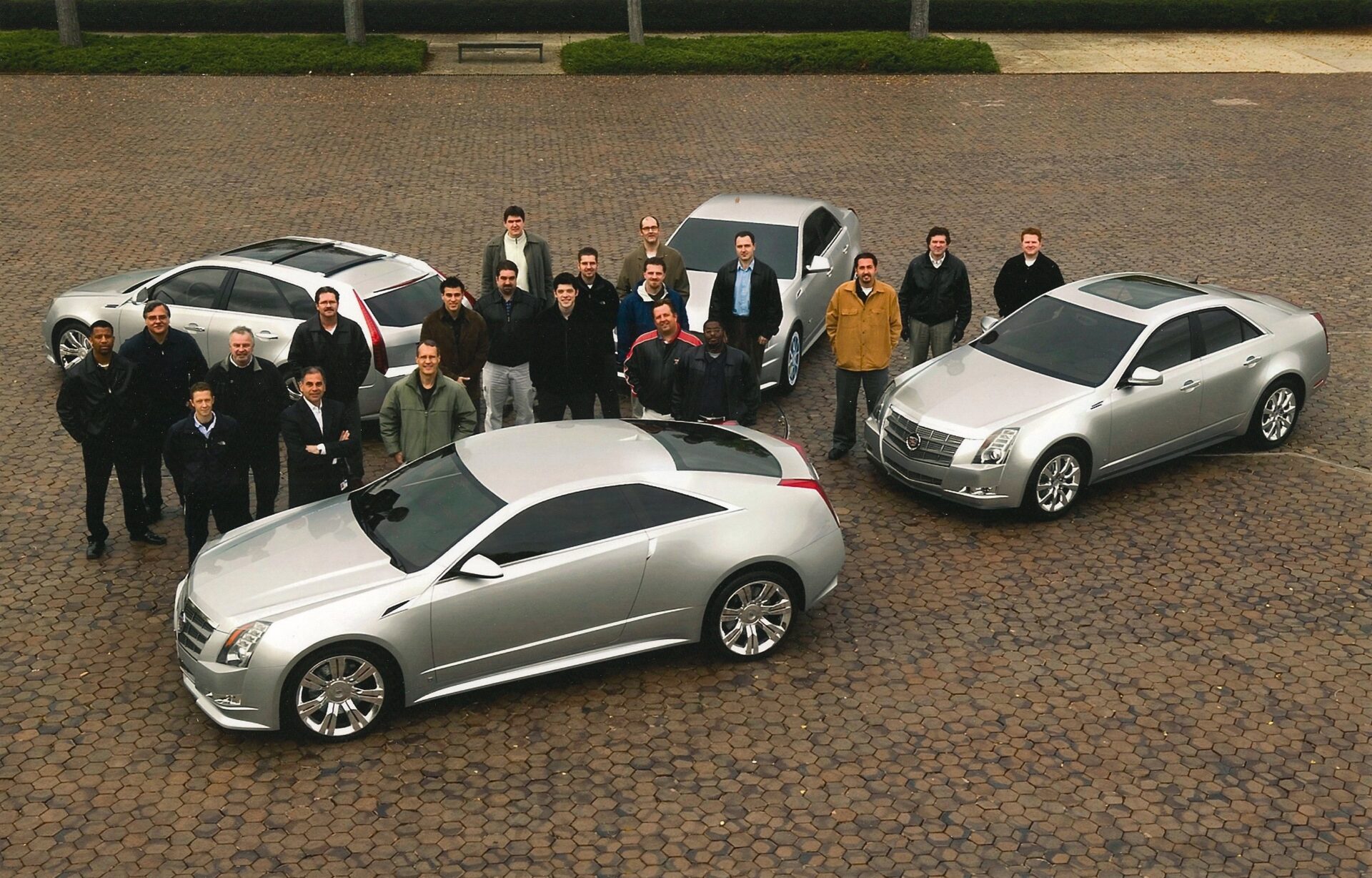 The Cadillac design team poses outdoors on a cobblestone plaza alongside three silver Cadillac CTS models: a Coupe in the foreground, a Wagon on the left, and a Sedan on the right.