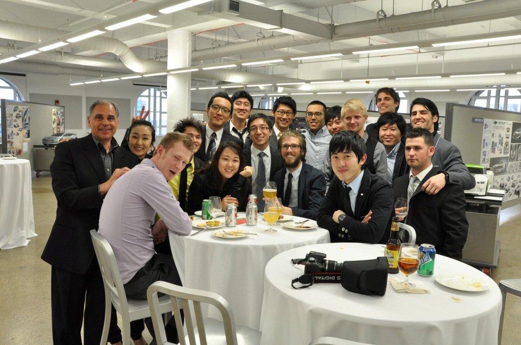 John Manoogian poses with a large group of smiling transportation design students gathered around a round table in a bright, modern studio room.