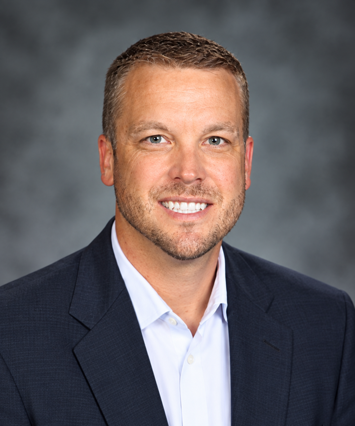 Professional headshot of a man with short brown hair and a smile, wearing a dark suit jacket and light collared shirt.