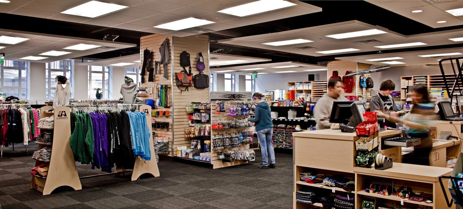 Wide view of a well-lit campus bookstore featuring racks of colorful apparel, display shelves with merchandise, and staff working behind the checkout counter.
