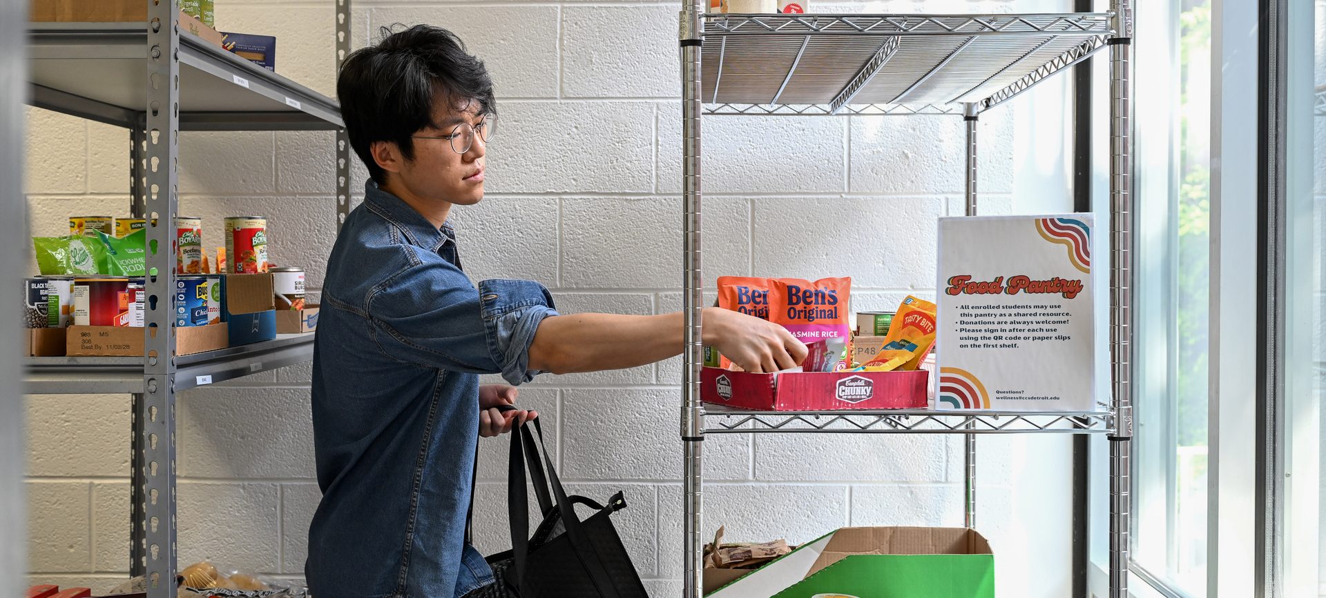 Young man in a denim shirt carrying a reusable tote bag as he reaches for packaged items on metal shelving in a food pantry.