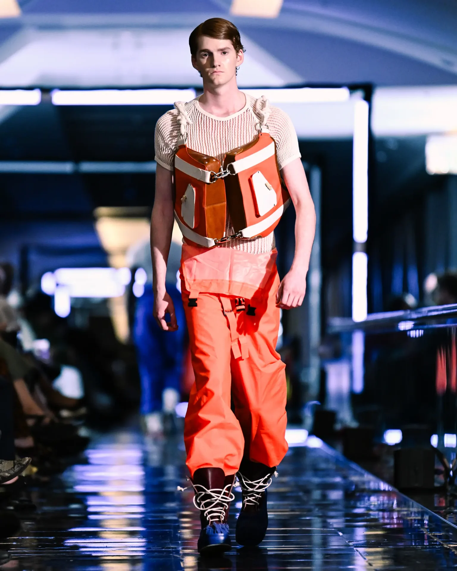 Model walking the runway wearing orange pants, a knit top and a orange leather vest.
