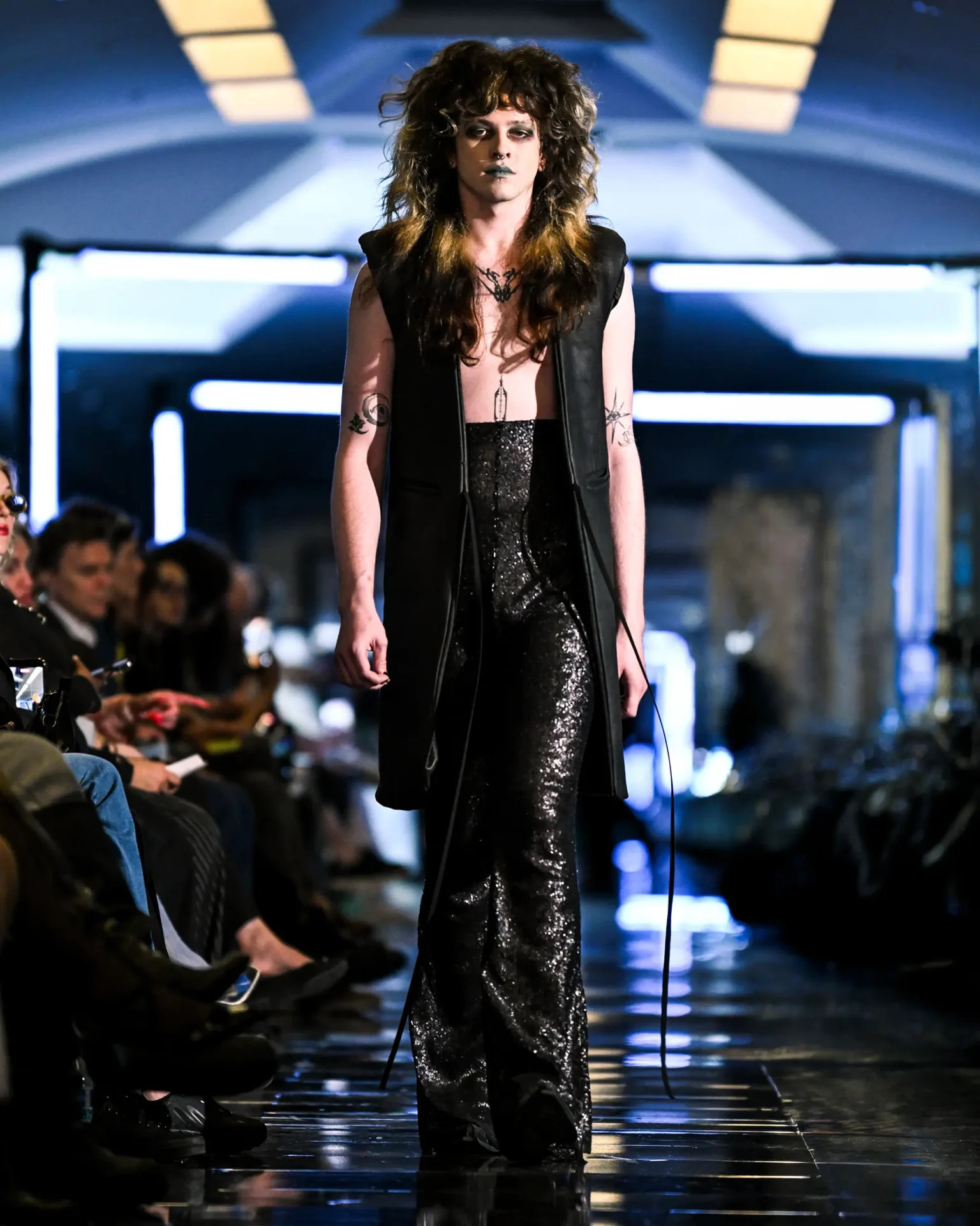 Model walking the runway wearing a black vest with sequined pants.