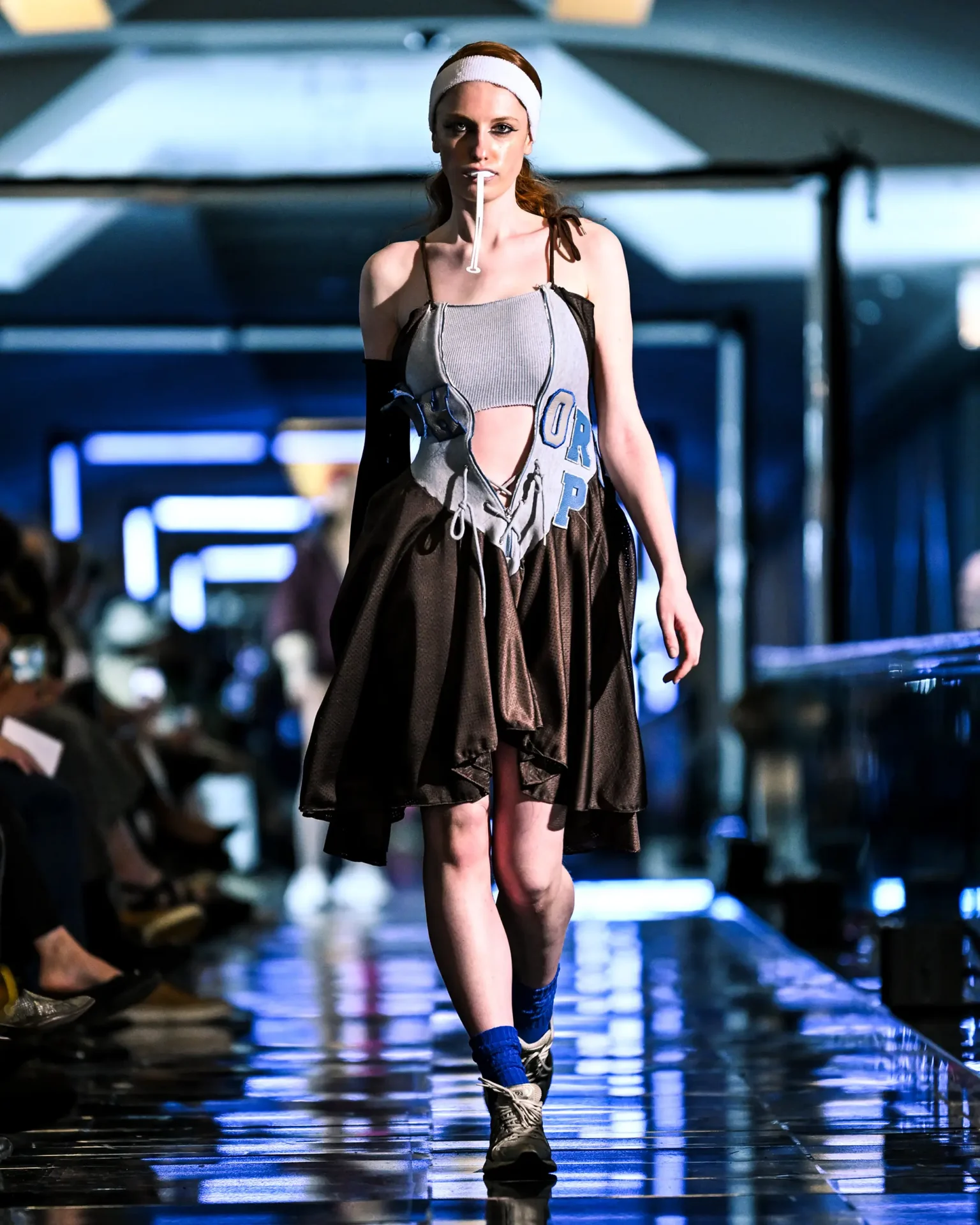 Model walking the runway wearing a gray and brown layered dress with blue lettering and athletic shoes.