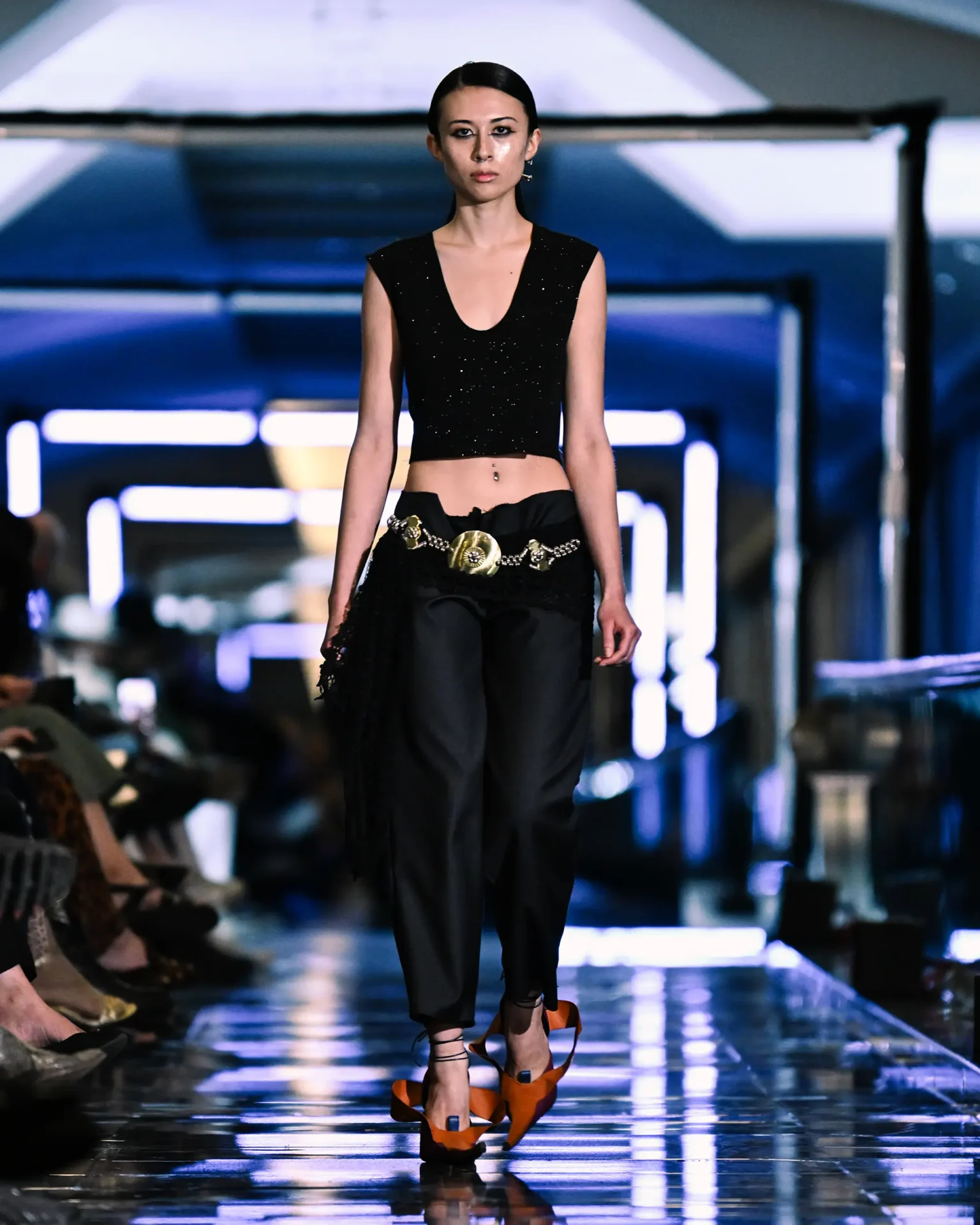 Model walking the runway wearing a black outfit with a decorative belt and orange shoes.