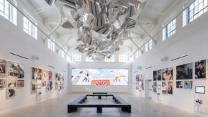 A bright, white-walled art gallery with high ceilings, featuring a large metallic sculpture suspended overhead, photo displays on the side walls, and a rectangular bench in the center.