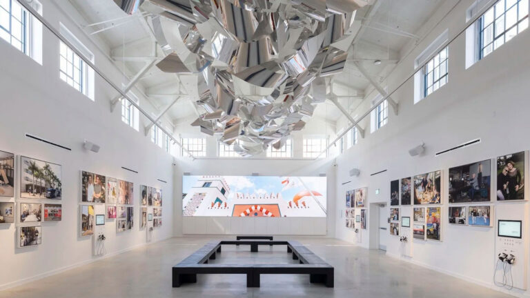 A bright, white-walled art gallery with high ceilings, featuring a large metallic sculpture suspended overhead, photo displays on the side walls, and a rectangular bench in the center.
