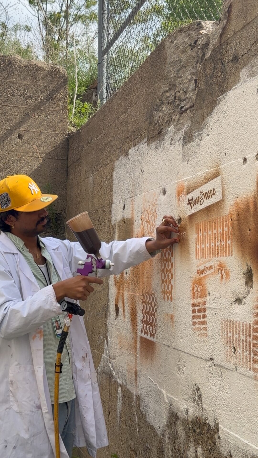 A person wearing a white lab coat and yellow baseball cap holding a pneumatic spray gun with dark liquid while placing a stencil onto a textured concrete outdoor wall covered in orange stenciled patterns and logos.