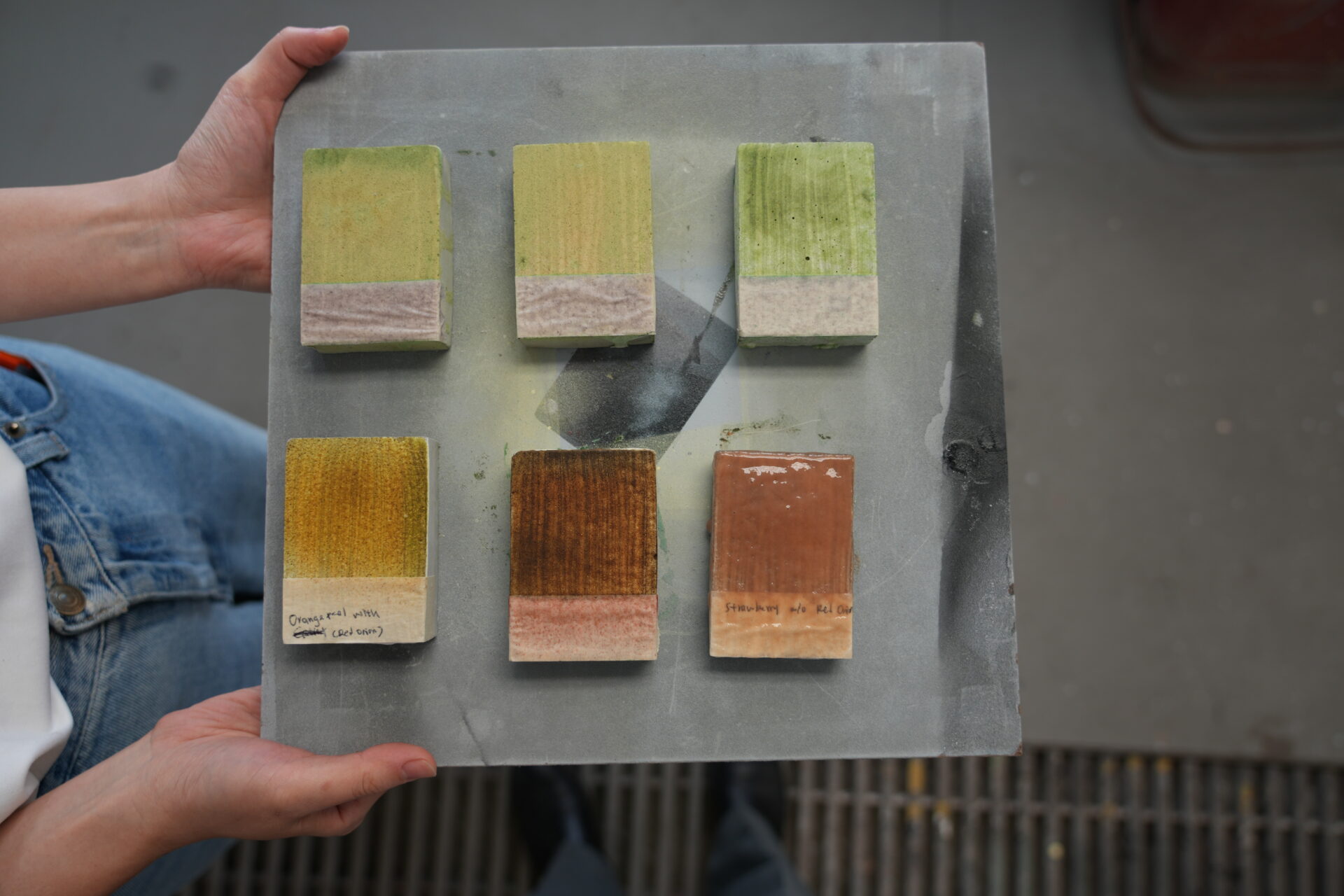 Hands holding a square board displaying six rectangular test blocks coated with brushed paint samples in shades of green, yellow, brown, and reddish-tan.
