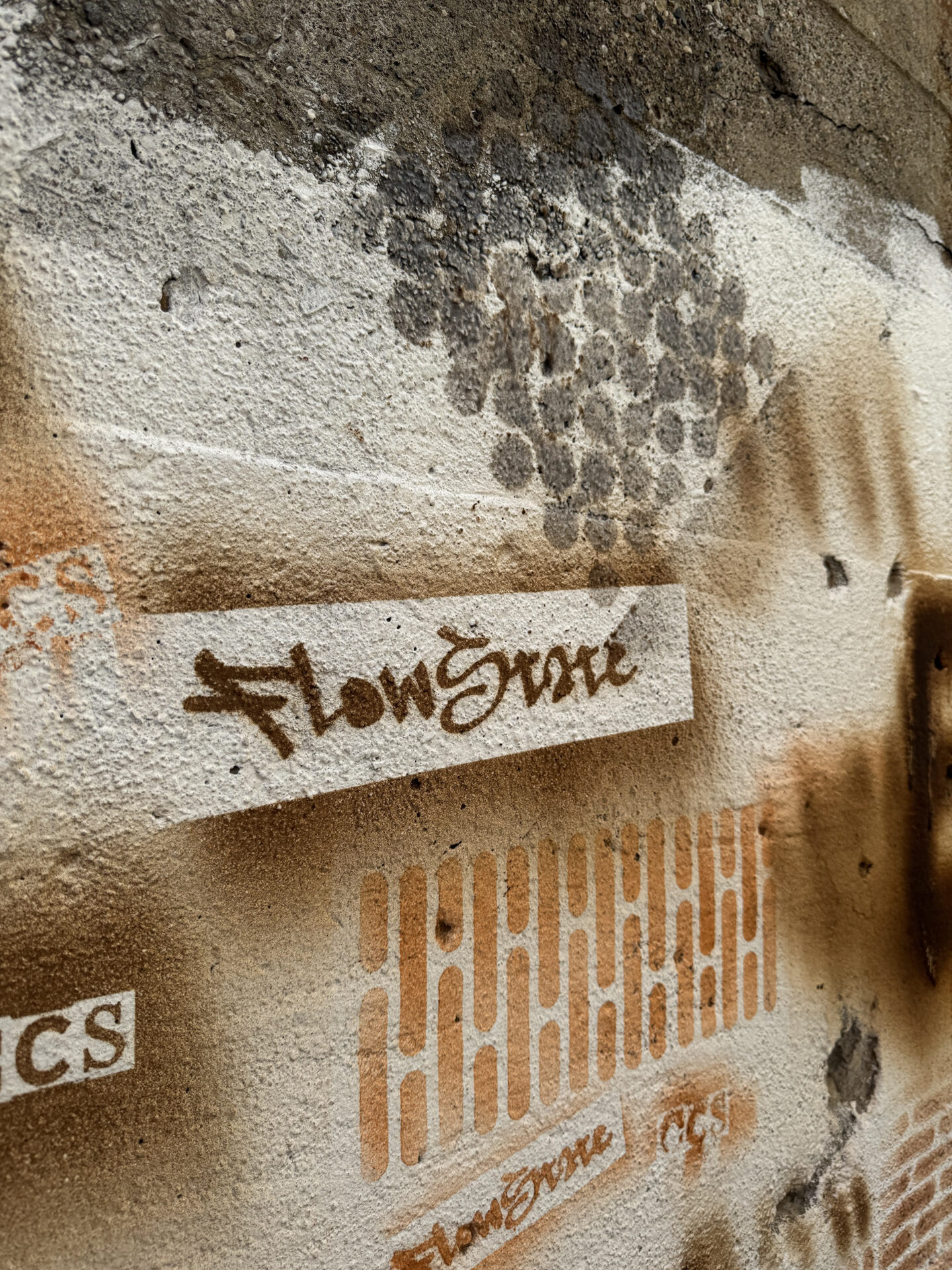 Close-up of a textured concrete wall displaying test-applied pigments using stencils, including stylized graffiti lettering for "FlowState" and geometric patterns in earth-toned brown and orange.