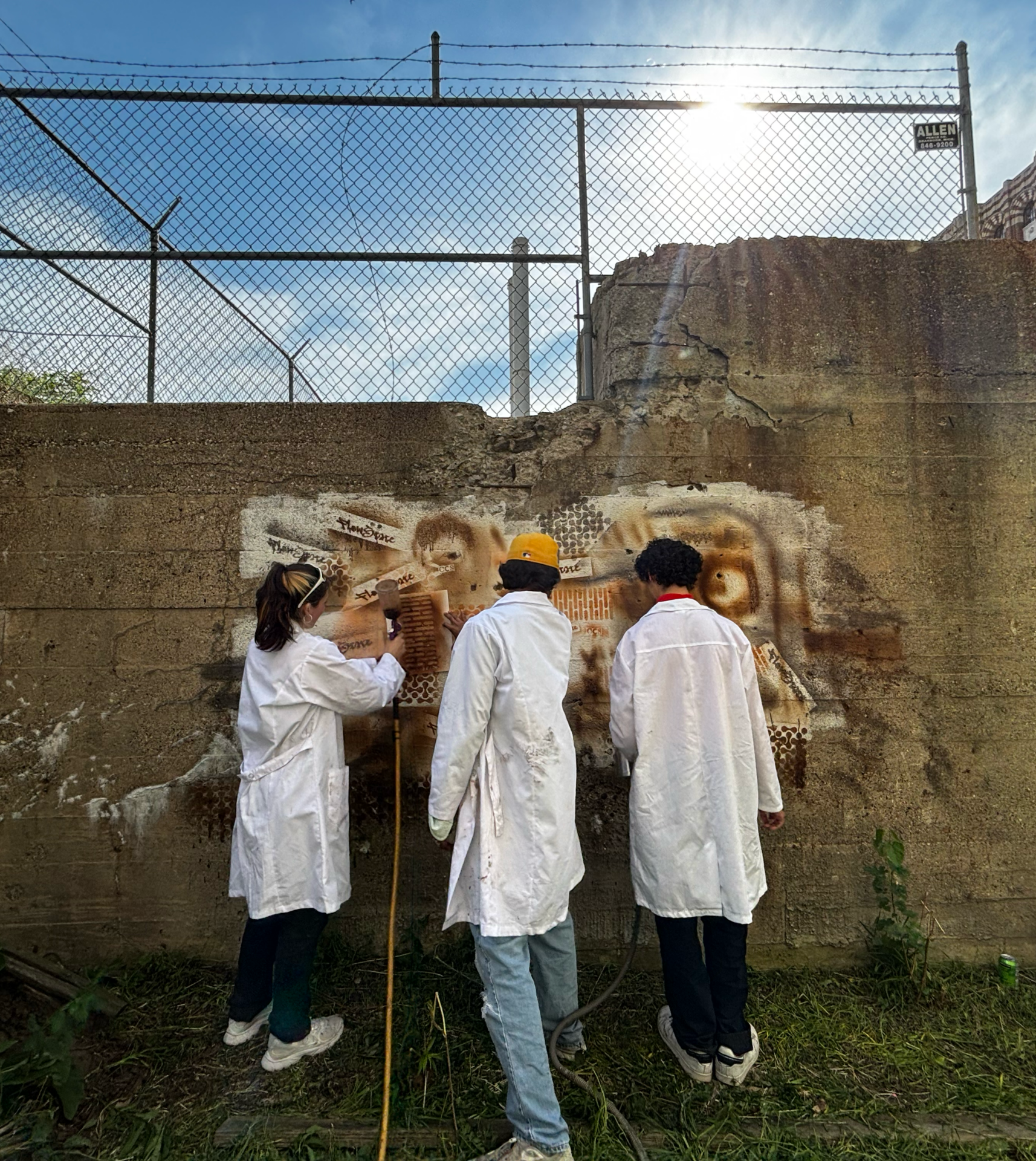 Three team members in white lab coats field-test spray paint against a tall concrete wall topped with a chain-link fence. One person uses a spray gun through a stencil while the others observe the application.