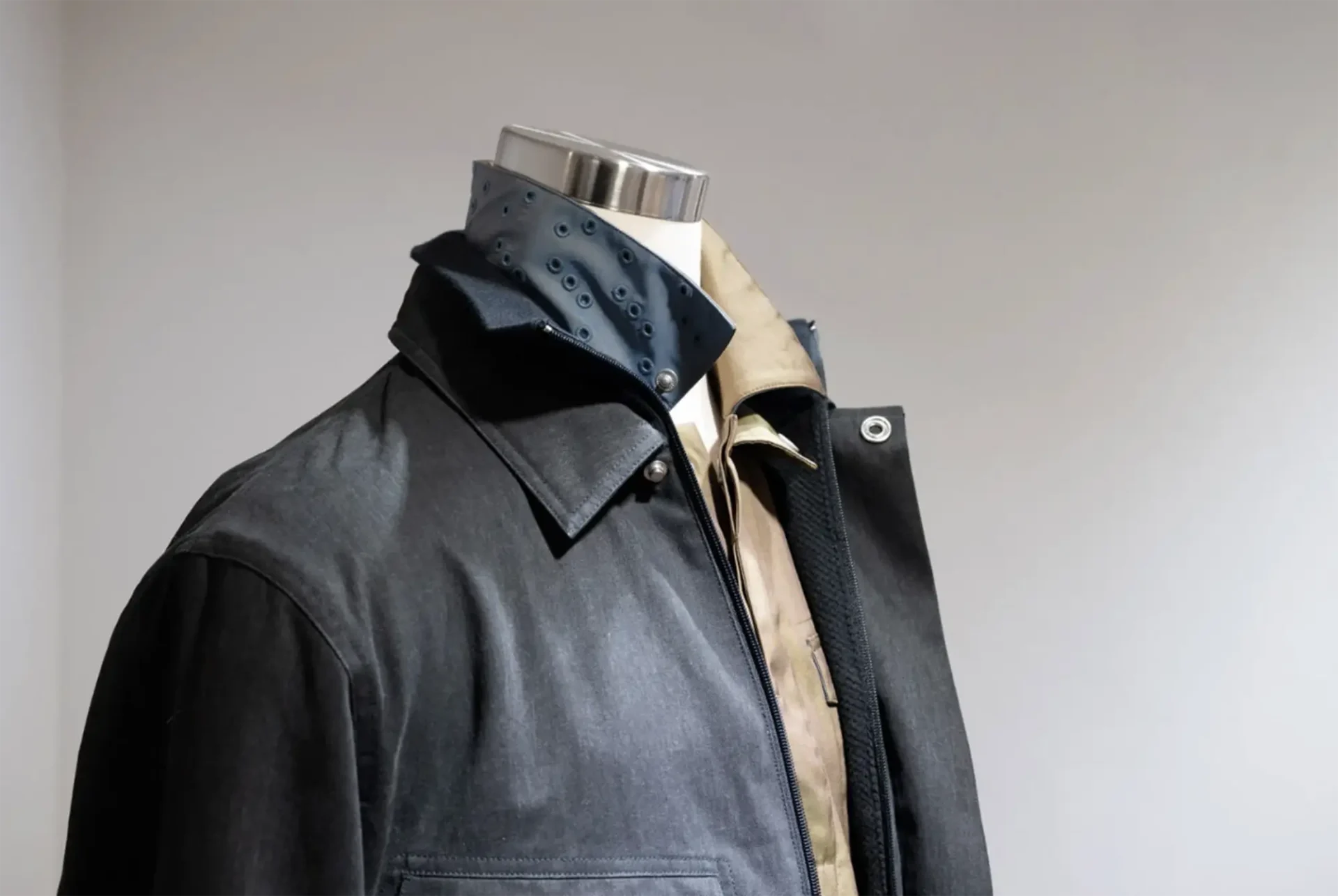 A mannequin displaying layered clothing, including a dark jacket, a brown collared shirt, and a dark blue inner collar detailed with rows of metal eyelets.